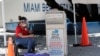 A poll worker wears personal protective equipment as she monitors a ballot drop box for mail-in ballots outside of a polling station during early voting, Aug. 7, 2020, in Miami Beach, Florida. 